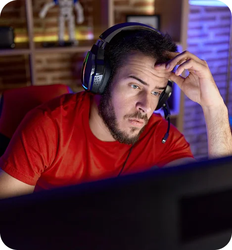 Skoncentrowany gracz w czerwonej koszulce z nausznikami, trzymający głowę podczas intensywnej gry online na ekranie komputera.