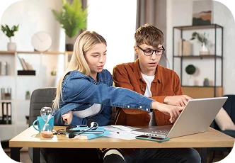 Dwóch studentów współpracuje w domu przy laptopie, omawiając zadania domowe i wspólnie pracując nad projektem naukowym.